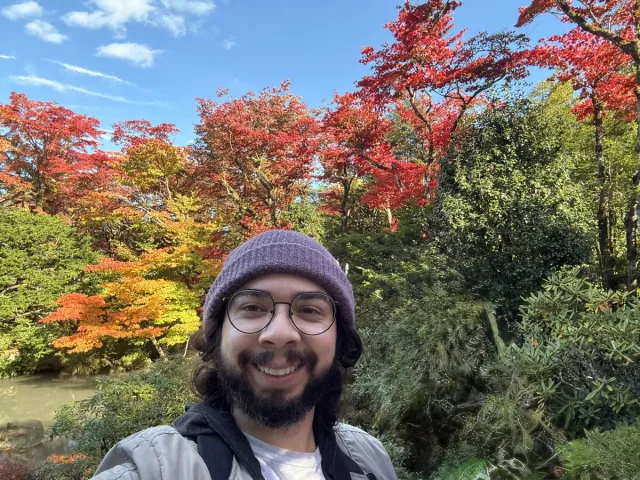 Bryan in a knit hat smiling in front of bright red and orange autumn trees